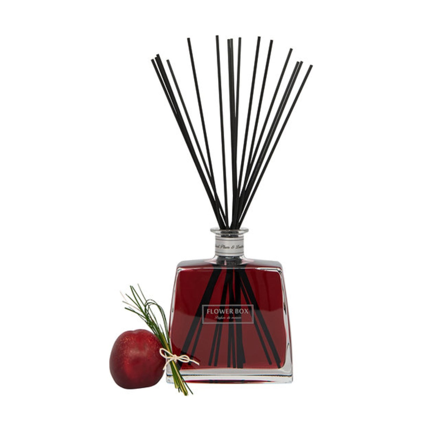 Image of a clear glass diffuser bottle with red liquid, square in shape, with a narrow neck and a light grey label around the neck. A bundle of long, thin grey reeds protrudes from the neck. In front of the bottle, on a white background, is a plum.