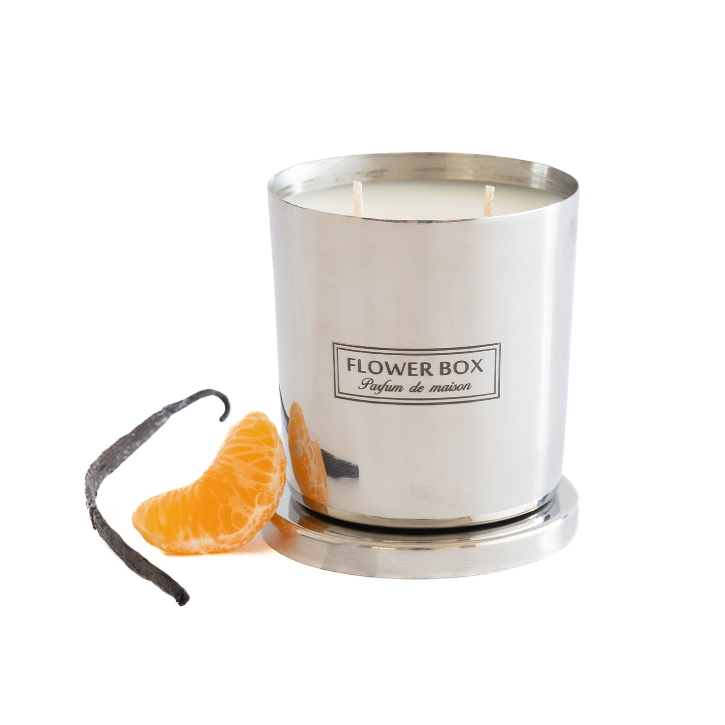 Image of a silver, brushed metal candle container with three wicks and a matching circular base. A rectangular label with black text on a white background reads "FLOWER BOX Pufum de maison". In front of the candle is a vanilla bean and a segment of a mandarin. The background is white