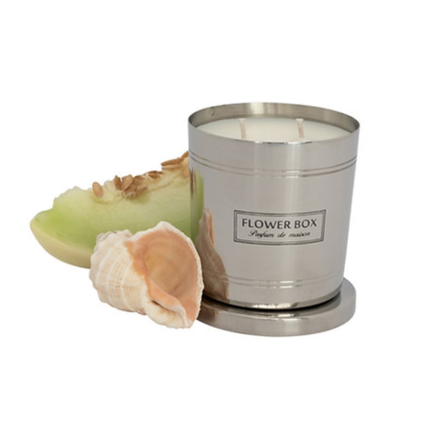 Image of a silver, brushed metal candle container with three wicks and a matching circular base. A rectangular label with black text on a white background reads "FLOWER BOX Pufum de maison". In front of the candle is a melon slice and shell. The background is white.