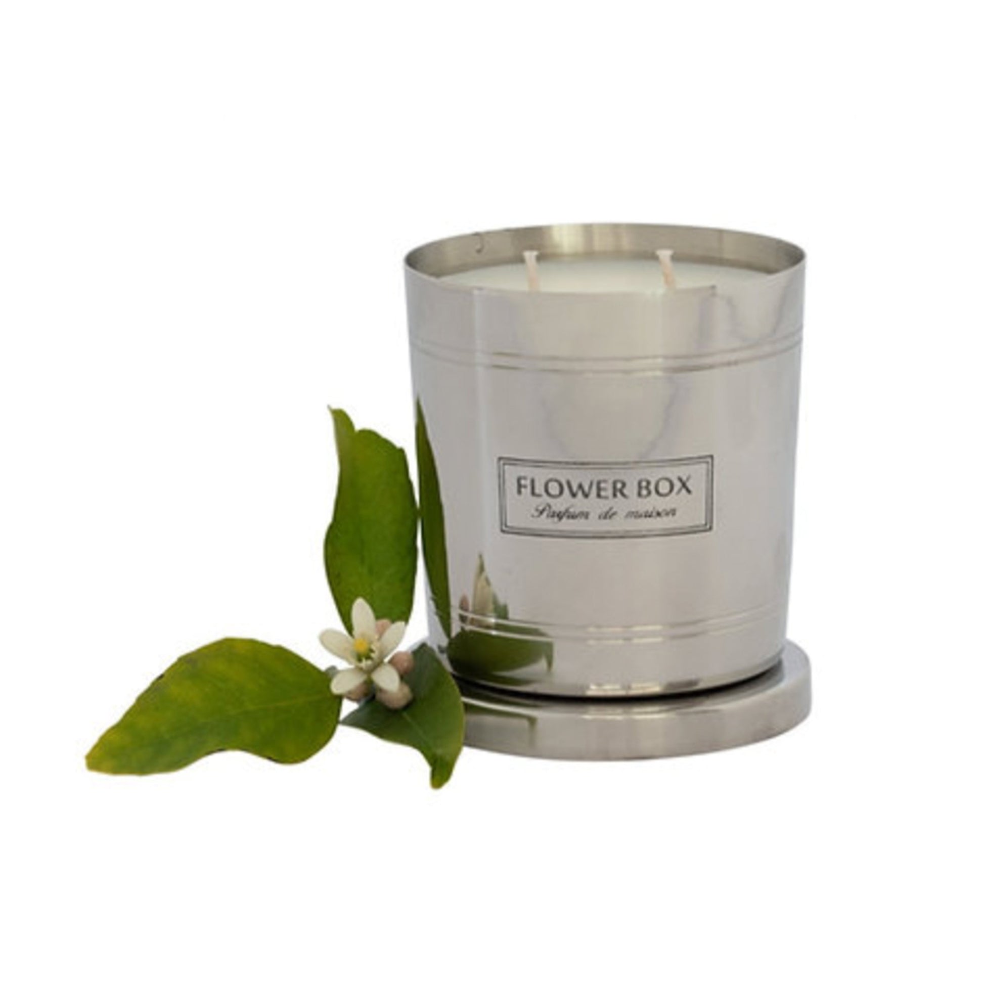 Image of a silver, brushed metal candle container with three wicks and a matching circular base. A rectangular label with black text on a white background reads "FLOWER BOX Pufum de maison". In front of the candle is a fig leaf. The background is white.