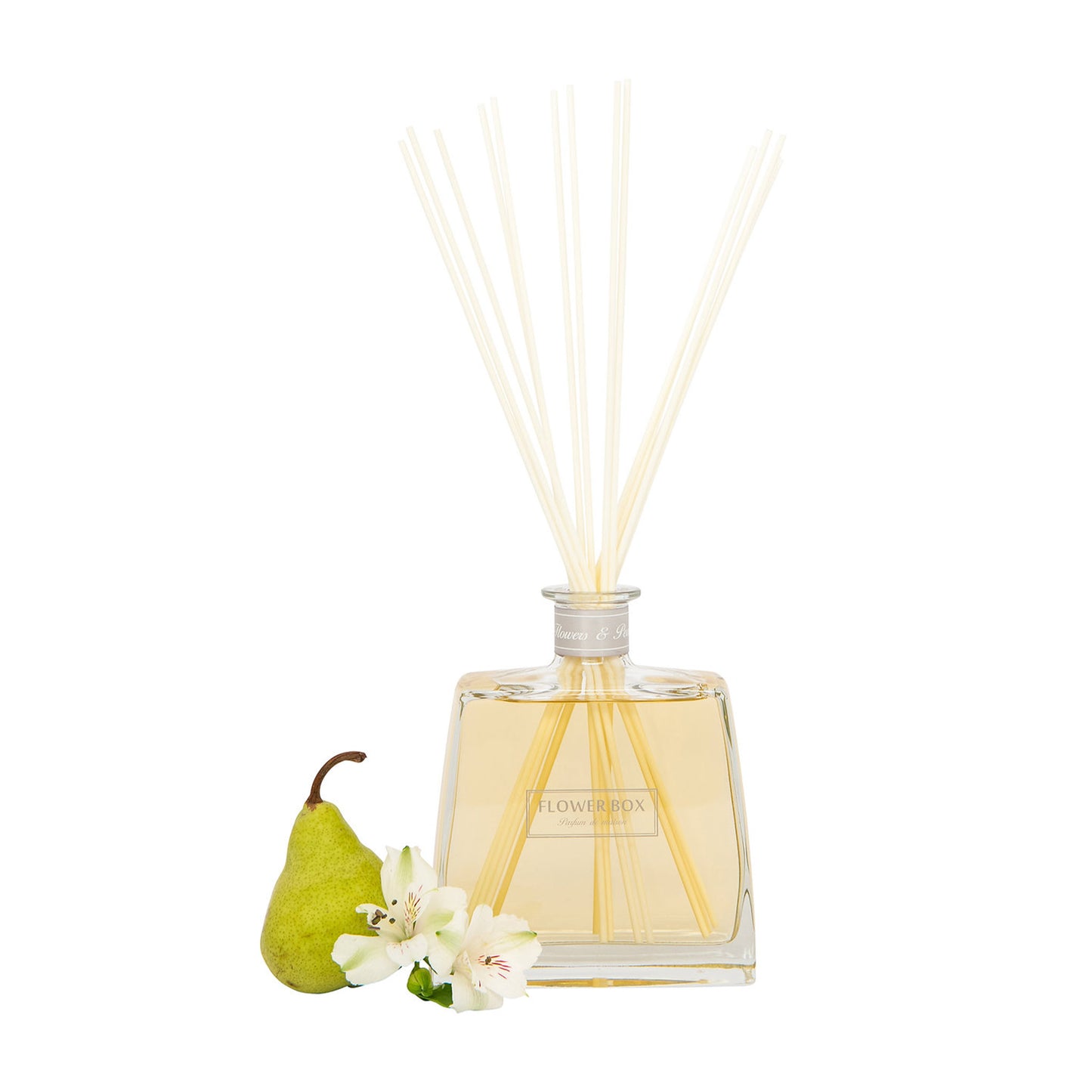 Image of a clear glass diffuser bottle, square in shape, with a narrow neck and a light grey label around the neck. A bundle of long, thin grey reeds protrudes from the neck. In front of the bottle, on a white background, is a pear and flowers