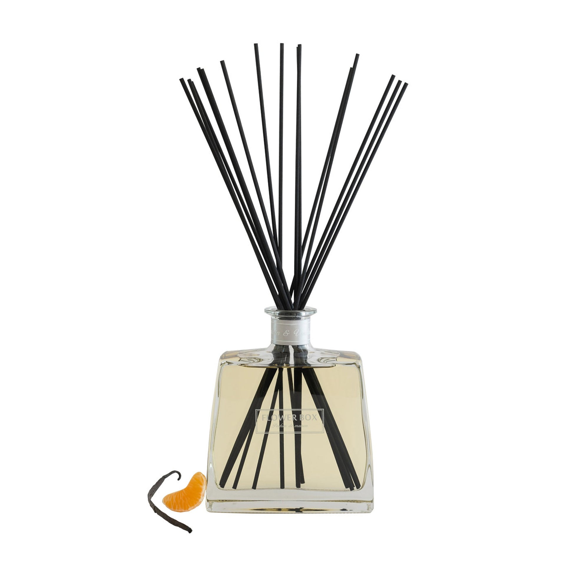 Image of a clear glass diffuser bottle, square in shape, with a narrow neck and a light grey label around the neck. A bundle of long, thin grey reeds protrudes from the neck. In front of the bottle, on a white background, is a vanilla bean and segment of a mandarin.