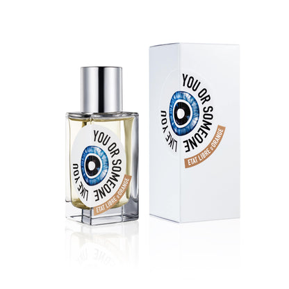 Image of Etat Libre d'Orange You Or Someone Like You Eau de Parfum in a clear rectangular glass perfume bottle with a silver spray nozzle cap. The bottle has a label with the words "You Or Someone Like You" and "Etat Libre d'Orange" and a graphic of a stylized blue eye with a black pupil. Behind the bottle is a white rectangular box with a similar label. Both are reflected on a white glossy surface.